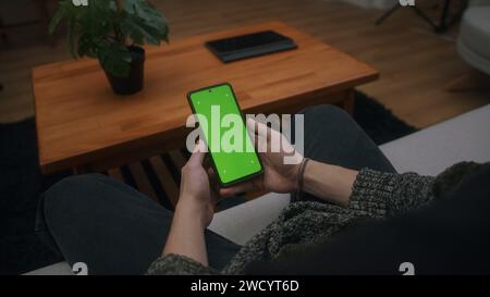 Young man using, tapping on screen smartphone with green mock-up screen. Male sitting on sofa, relaxing at home. Close-up over the shoulder shot Stock Photo