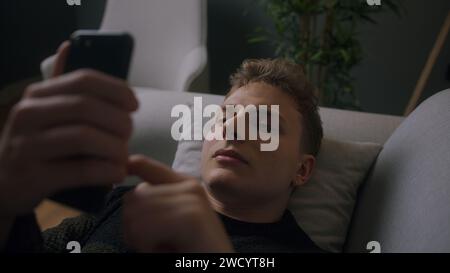 Close-up young blonde man uses smartphone while laying on a sofa at home at night. Man browses through internet, watches videos and uses social networ Stock Photo