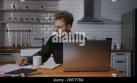 Blonde young university student sitting in the kitchen at home, looking laptop computer, studying, taking notes. Male student researching, study at ho Stock Photo