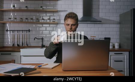 Young university student sitting in the kitchen at home, looking laptop computer, studying, taking notes. Male student researching, study at home Stock Photo