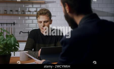 Two co-worker men are sitting in the kitchen having a fun chat and laughing. Taking a break at work, relaxing time. OTS shot Stock Photo