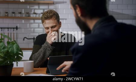 Two young co-worker men are sitting in the kitchen having a fun chat and laughing. Taking a break at work, technology company workers, relaxing time. Stock Photo
