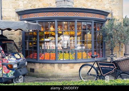 Fruit and vegetables and an old fashioned weighing scale in the window of the Broadway Deli, with a delivery bicycle outside. Broadway, England Stock Photo