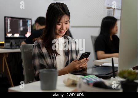 Young beautiful Asian female graphic designer is in the office, using her smartphone at her office desk. Business people and creative job concepts Stock Photo