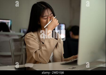 A tired and overworked young Asian businesswoman or female office employee is suffering from eye strain while working on her project on computer at he Stock Photo