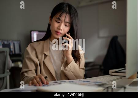 A professional Asian businessman is talking on the phone with her supplier, having a serious phone call, working at her desk in the office. Stock Photo