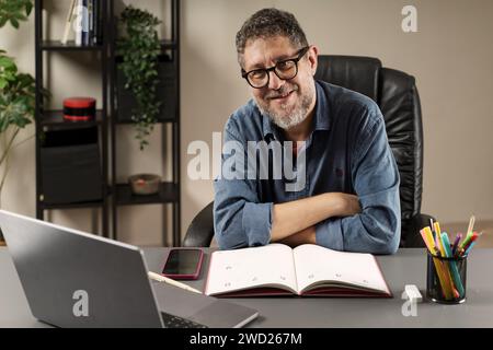 Cheerful businessman in casual attire working at an organized desk with a laptop and planner. Stock Photo