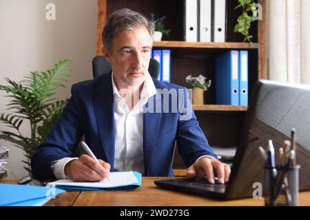 Businessman working in the home office on a laptop writing on some documents on table with shelves in the background Stock Photo