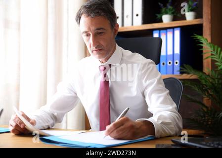 Man in white shirt and red tie writing on a document working in an office with wooden furniture Stock Photo