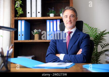 Smiling businessman with arms crossed in his home office wearing blue suit and documents on wooden table and bookshelves background. Stock Photo