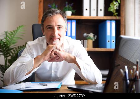 Man in white shirt at work smiling leaning on an office table looking ahead with documents and laptop and shelves with folders in the background Stock Photo