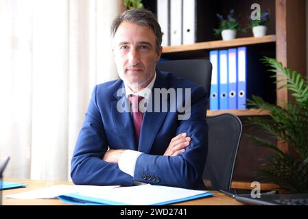 Businessman in his home office wearing blue suit and red tie looking straight ahead with documents on wooden table and bookshelves background. Stock Photo