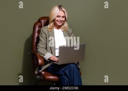 Photo of smm agency worker mature woman sit home office leather armchair doing daily tasks using laptop isolated on khaki color background Stock Photo