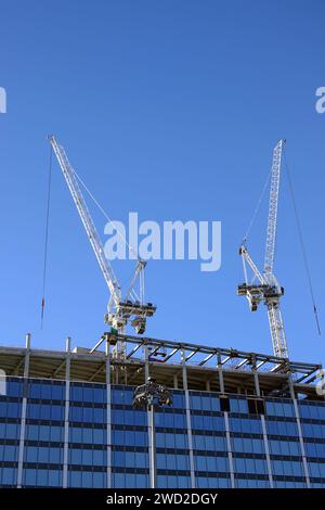 Two hoisting tower cranes on the roof of being construction of modern high skyscraper  building against blue cloudless sky Stock Photo