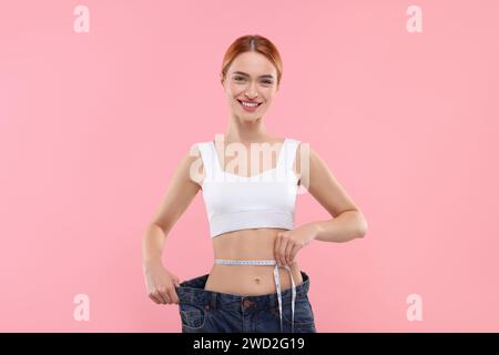 Slim woman wearing big jeans and measuring waist with tape on pink background. Weight loss Stock Photo