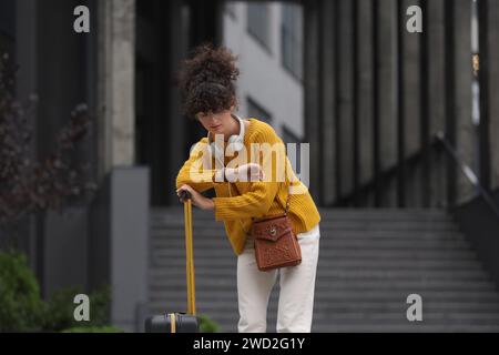 Being late. Worried woman with suitcase looking at watch near building outdoors Stock Photo