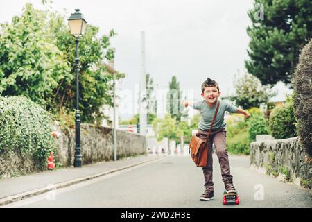 Outdoor portrait of funny little schoolboy wearing brown leather bag over shoulder, riding skateboard. Back to school concept. Film look toned image Stock Photo