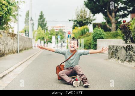 Outdoor portrait of funny little schoolboy wearing brown leather bag over shoulder, riding skateboard. Back to school concept. Film look toned image Stock Photo