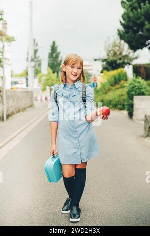Outdoor portrait of funny little schoolgirl, wearing formal dress and backpack, holding lunch box and red apple. Back to school concept, film look ton Stock Photo