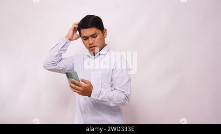Asian man scratch his head while holding mobile phone and showing confused face expression against white background Stock Photo