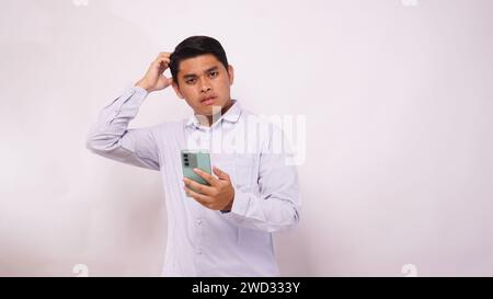Asian man scratch his head while holding mobile phone and showing confused face expression against white background Stock Photo