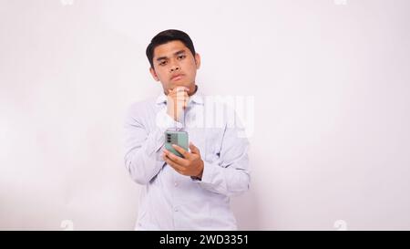 Asian man scratch his head while holding mobile phone and showing confused face expression against white background Stock Photo