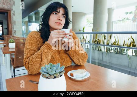 young Latin Venezuelan woman with long black hair, sitting alone in cafeteria drinking coffee and eating pudding alone and thoughtful, relaxed, enjoyi Stock Photo