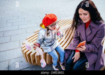 Brunette caucasian positive woman and daughter using smartphone sitting on the bench at city street. Mother calms the girl showing cartoon on the Stock Photo