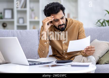 Concerned young Indian man sitting on a couch in home setting, examining papers with a laptop and calculator nearby. Stock Photo