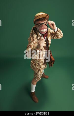 Funny image of young man in image of detective looking in magnifying glass against green studio background. Big teeth Stock Photo