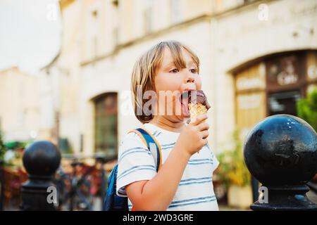 Cute little boy eating chocolate ice cream outdoors, wearing backpack, travel with kids Stock Photo