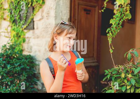 Cute little girl eating blue ice cream outdoors, wearing orange shirt and backpack, travel with kids Stock Photo