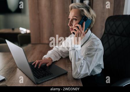 Side view of busy middle-aged businesswoman with blonde hair talking on smartphone siting at office desk with laptop, share helpful information Stock Photo