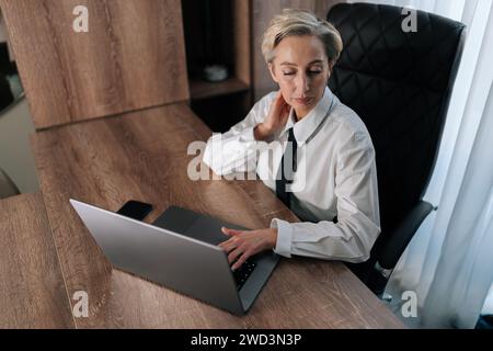 Exhausted middle-aged business woman having neck pain, pinched nerves, tensed muscles, cervical osteochondrosis. High-angle view of tired female Stock Photo