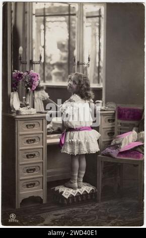 An antique postcard shows a small beautiful girl in a white dress, standing in front of the dressing table, half turned towards the camera. Bavaria, G Stock Photo