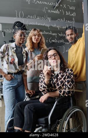 Woman with disability writing code on glass wall in office, her colleagues standing behind her Stock Photo