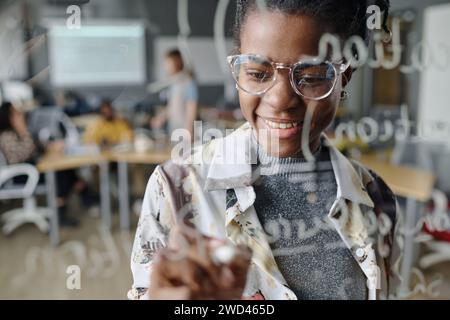 Cheerful woman making notes with marker on glass wall in office, her coworkers sitting on background Stock Photo