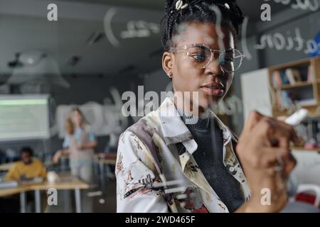 Female coder thoughtfully writing on glass wall in office, her colleagues working on background Stock Photo