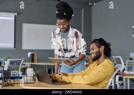 Cheerful indian programmer working on laptop at table in office, his black colleague standing nearby Stock Photo