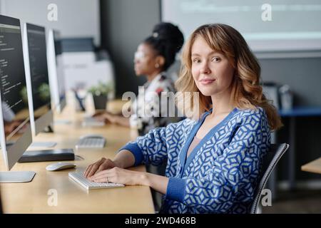 Caucasian female coder sitting at her desk working with data on computer and smiling at camera Stock Photo