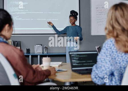 Black female office employee pointing on digital screen with codes, her female coworkers listening Stock Photo