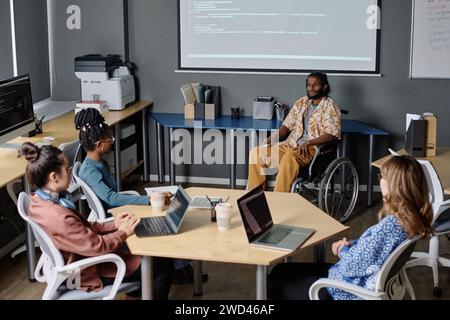 Indian man in wheelchair giving speech to his colleagues in office, code projected on digital board Stock Photo