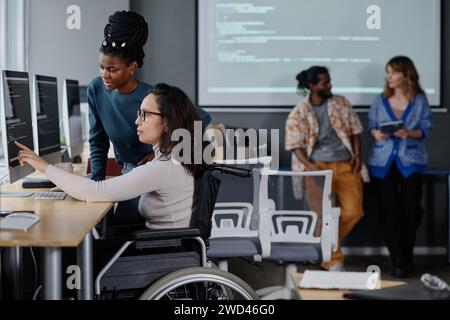 Black woman helping her colleague in wheelchair with code writing, coworkers standing on background Stock Photo