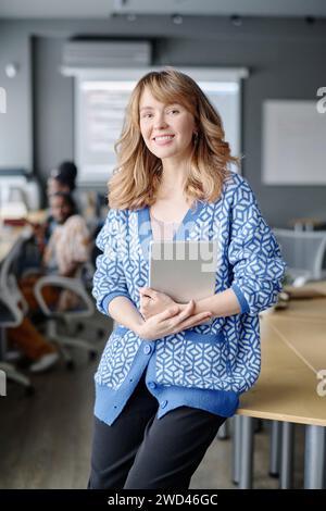 Cheerful caucasian woman posing in office with tablet in hands, her coworkers sitting on background Stock Photo