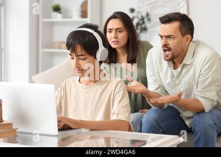 Teenage boy in headphones using laptop and his angry parents at home. Family problem concept Stock Photo