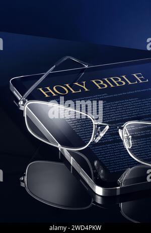 This close-up image shows a tablet, either Android or iOS, with the words Holy Bible and a passage from the Bible written on it. A pair of reading gla Stock Photo