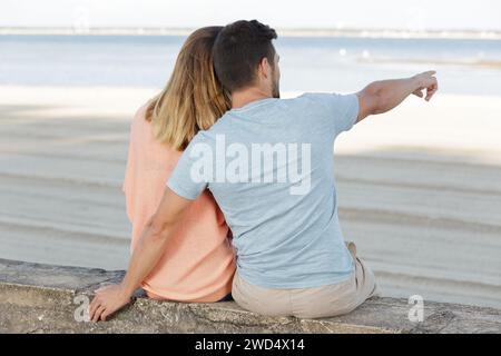 couple sat on wall overlooking beach and pointing into distance Stock Photo