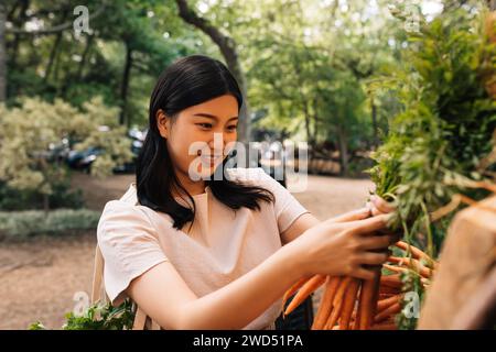 Young smiling customer choosing carrot on the outdoor market. Woman holding fresh vegetables while standing outdoors. Stock Photo