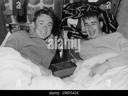 An antique photo with shows two young men lie on their beds and smile happily behind them, a suitcase and black shoes are visible. Germany. Date unkno Stock Photo