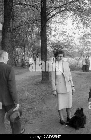 An antique photo with shows a woman in a shabby suit with a dog near her feet posing in the park, an elderly man in a formal suit walks past them, he Stock Photo